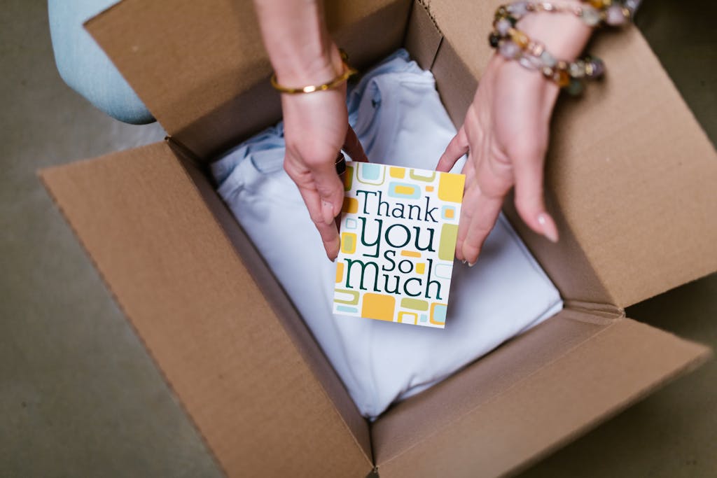 A person holds a thank you card above items in an open shipping box, suggesting care in sending.