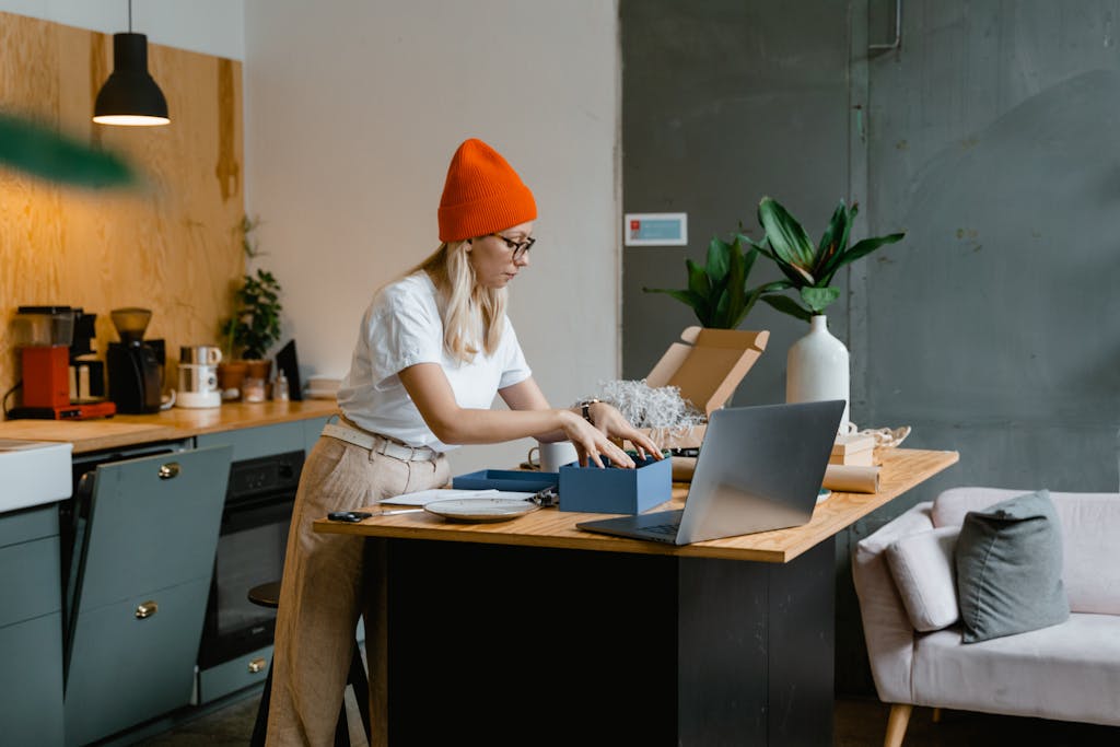 A young woman unpacks online shopping orders in a stylish modern kitchen, using her laptop for work.